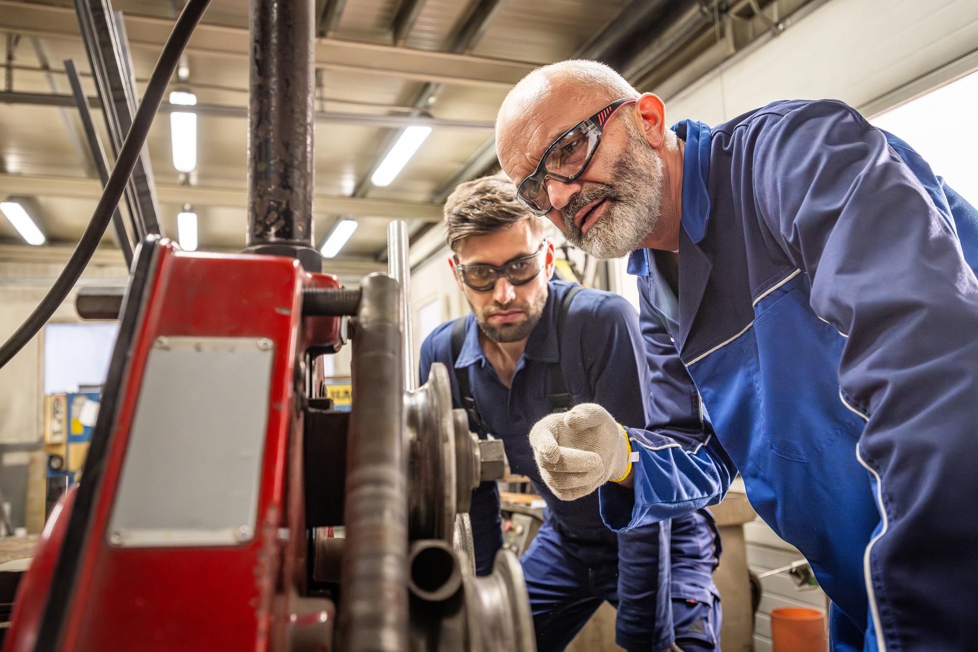 Two focused Caucasian males in protective gear use machinery to bend metal parts, collaborating efficiently in an industrial setting.