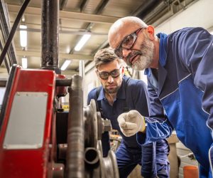 Two focused Caucasian males in protective gear use machinery to bend metal parts, collaborating efficiently in an industrial setting.